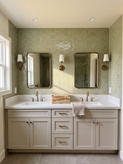 A modern bathroom vanity featuring a double sink with a white countertop, light beige cabinets, two rectangular mirrors, and a light sage green hexagonal tile backsplash.
