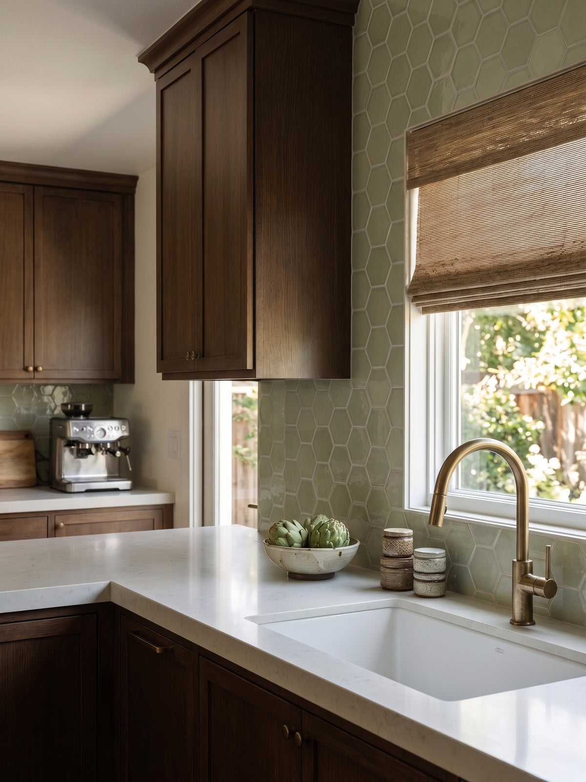 A modern kitchen corner with dark wood cabinets, a white countertop, a white double sink, a brass faucet, and a light green hexagonal tile backsplash. A window with a brown roller shade is on the right, and a coffee machine sits on the counter to the left.