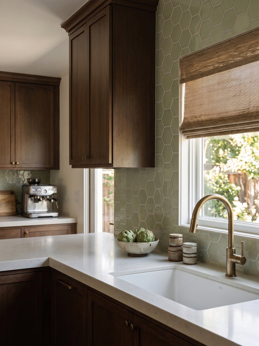 A modern kitchen corner with dark wood cabinets, a white countertop, a white double sink, a brass faucet, and a light green hexagonal tile backsplash. A window with a brown roller shade is on the right, and a coffee machine sits on the counter to the left.