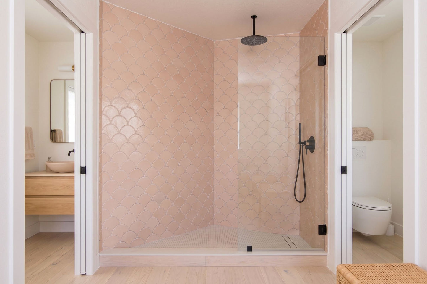 A modern bathroom interior featuring a walk-in shower with pale blush pink scalloped tile walls and clear glass doors, flanked by open doorways showing a vanity on the left and a toilet on the right.