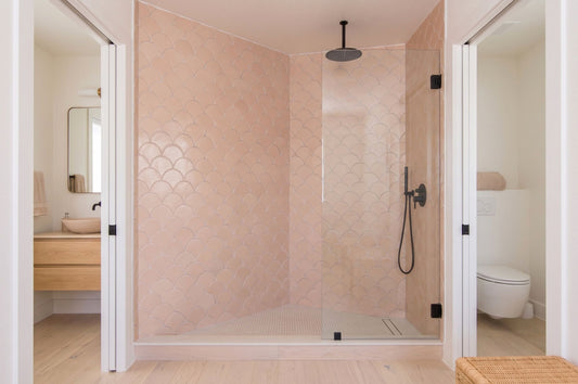 A modern bathroom interior featuring a walk-in shower with pale blush pink scalloped tile walls and clear glass doors, flanked by open doorways showing a vanity on the left and a toilet on the right.