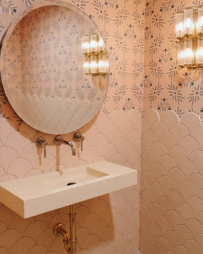 A modern bathroom corner featuring a white wall-mounted sink, a large round mirror, and warm-toned patterned wallpaper with scalloped tile detailing on the lower wall. Gold-toned wall sconces are turned on, casting a warm glow.