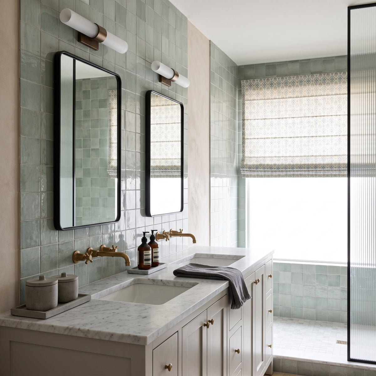 A modern bathroom vanity with a white marble countertop, double sinks, brass faucets, two black-framed mirrors, and pale green tiled walls. A window with a woven Roman blind is visible on the right, along with a glass shower screen.