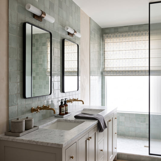 A modern bathroom vanity with a white marble countertop, double sinks, brass faucets, two black-framed mirrors, and pale green tiled walls. A window with a woven Roman blind is visible on the right, along with a glass shower screen.