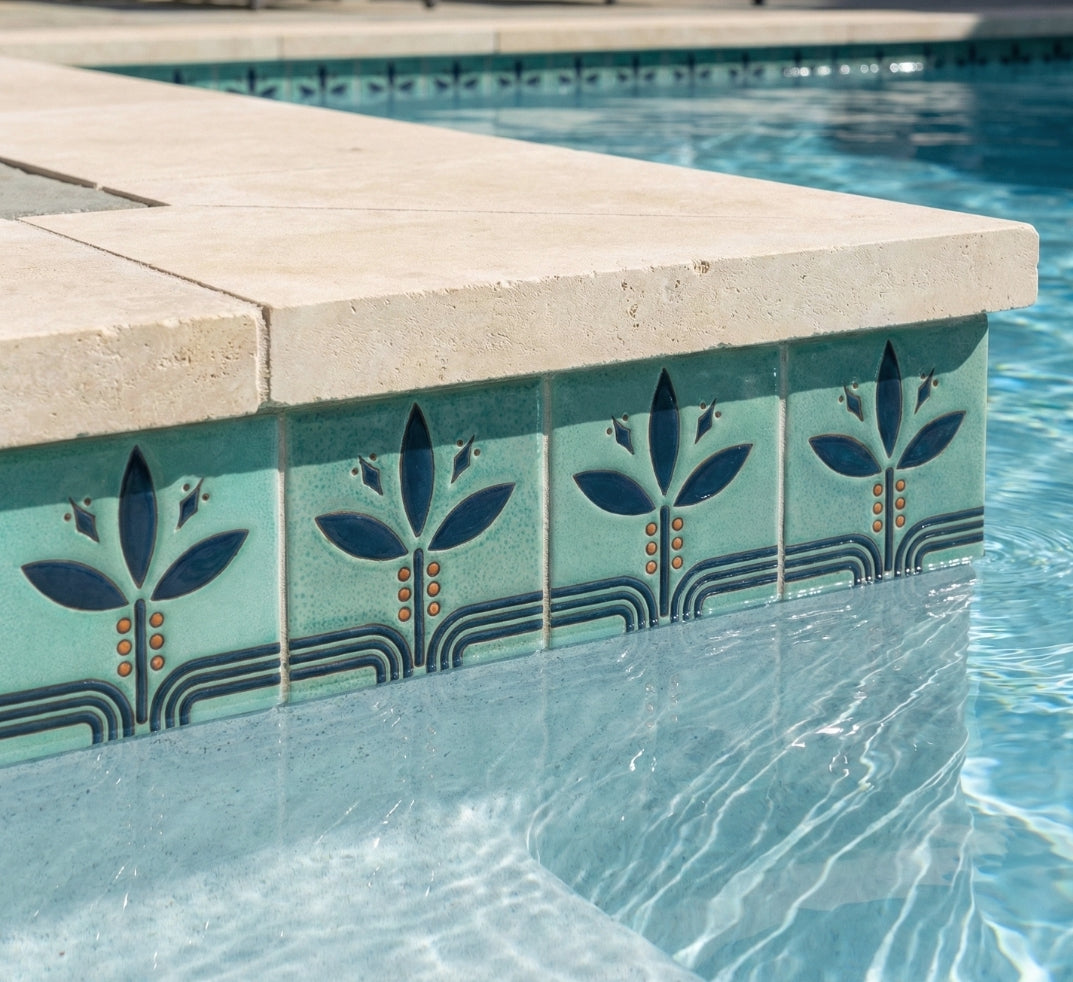Relief Bayas Jade Pool Waterline Tile by Clay Imports, featuring stylized dark blue leaf motifs, orange accents, and curved lines below a stone coping.