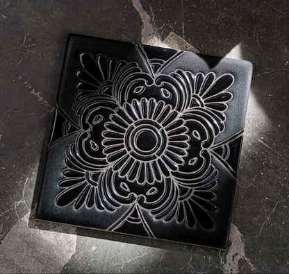 Black 6x6 relief tile with intricate floral embossing, matte finish, displayed on dark stone surface, top-down view