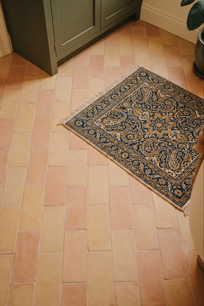 A small rectangular area rug with a navy blue and gold paisley pattern lies on a terracotta tile floor in a room corner near a dark green cabinet.