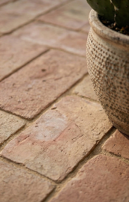 Terracotta rustic brick paver from Clay Imports Pavers, with an earthy textured surface and sandy grout, beside a decorative pot.