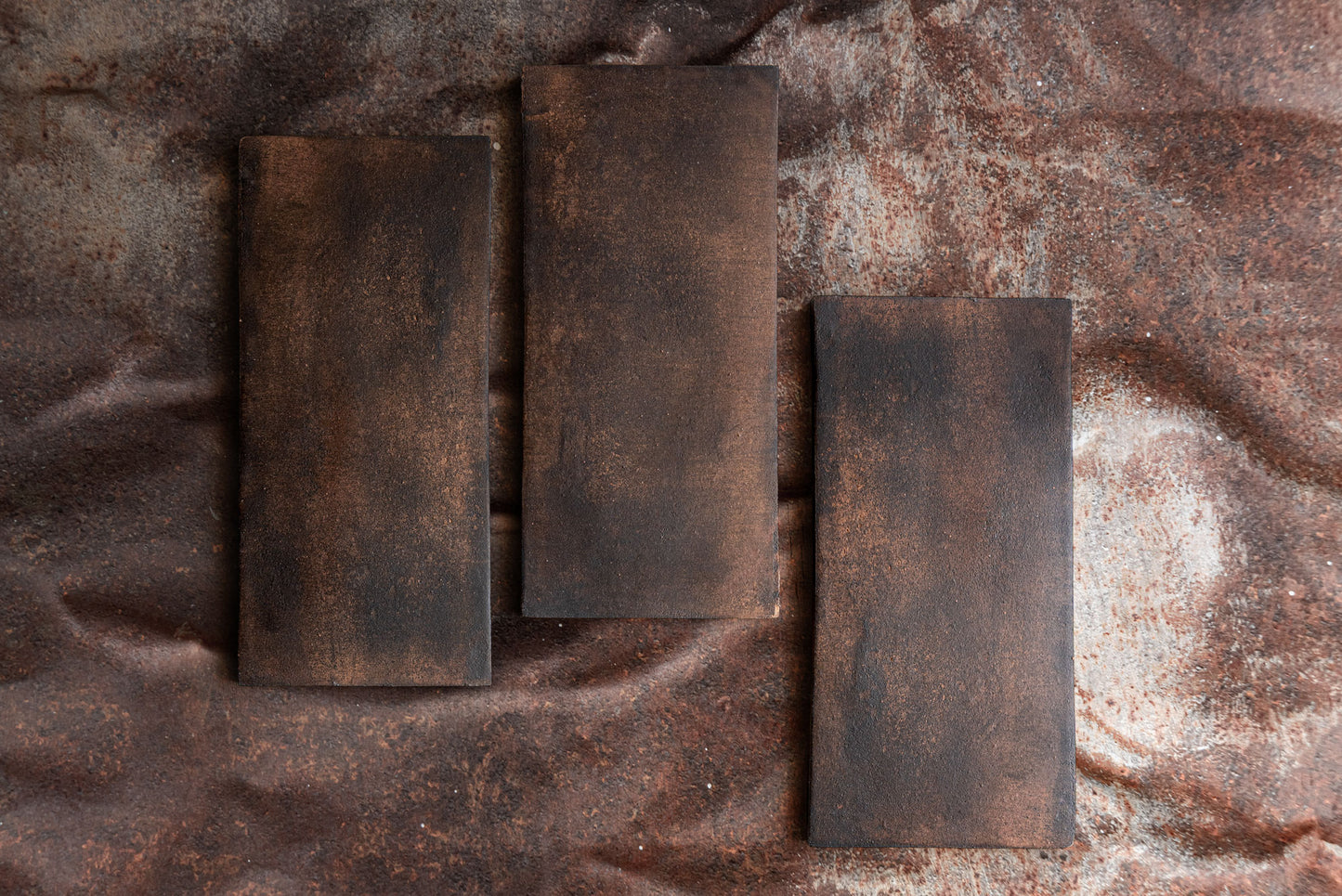 Dark brown rectangular Cotto Oxido 6x13 tiles with rustic, weathered texture arranged on a corroded metal surface, top-down view.