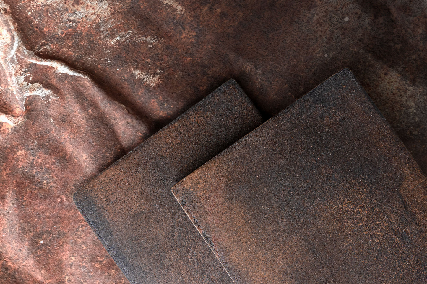 Rustic dark brown ceramic tile with textured oxidized finish, by Cotto, displayed in overlapping arrangement on weathered metal background
