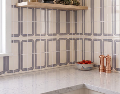 A kitchen backsplash featuring cream tiles with a gray geometric border pattern, paired with a white marble countertop and a light wood floating shelf.