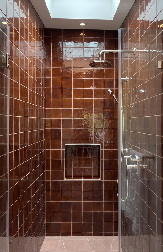 A modern walk-in shower enclosure with glossy brown rectangular wall tiles, a glass door, a built-in niche shelf, and a round shower head mounted on a chrome arm.