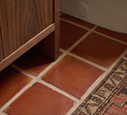 Terracotta tiles on a floor with a wooden cabinet and vintage rug corner.