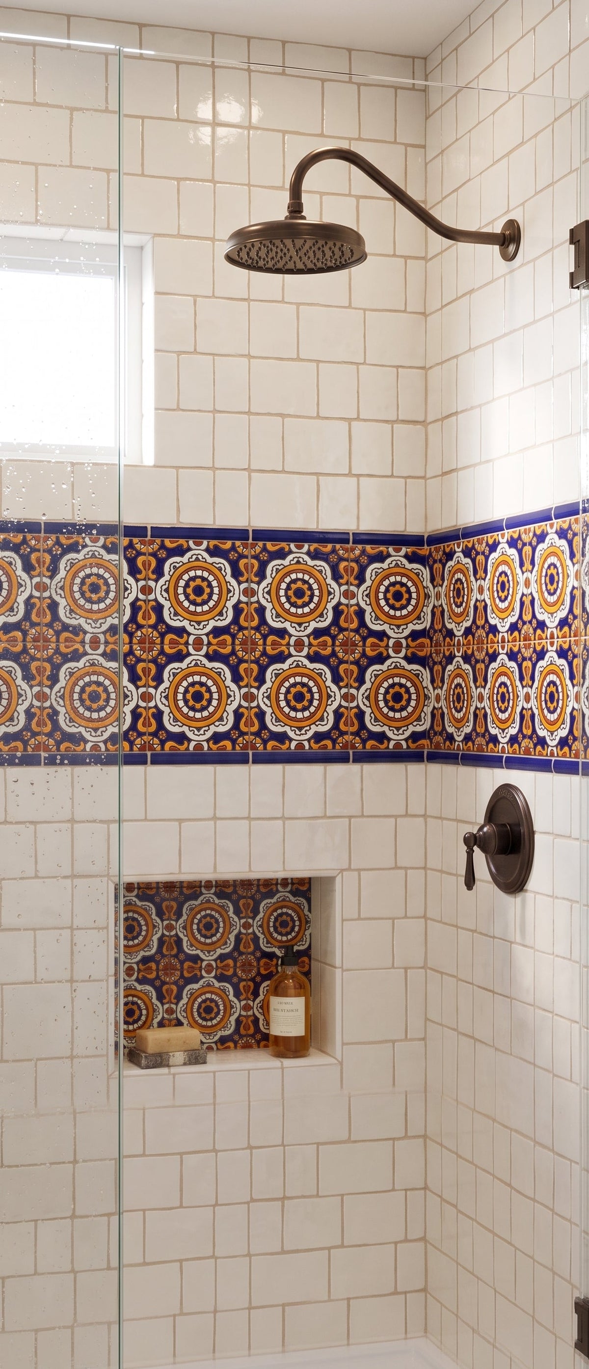 A walk-in shower features white subway tile walls accented by a vibrant band of blue, orange, and yellow patterned Spanish tiles, complemented by bronze fixtures and a matching built-in niche.