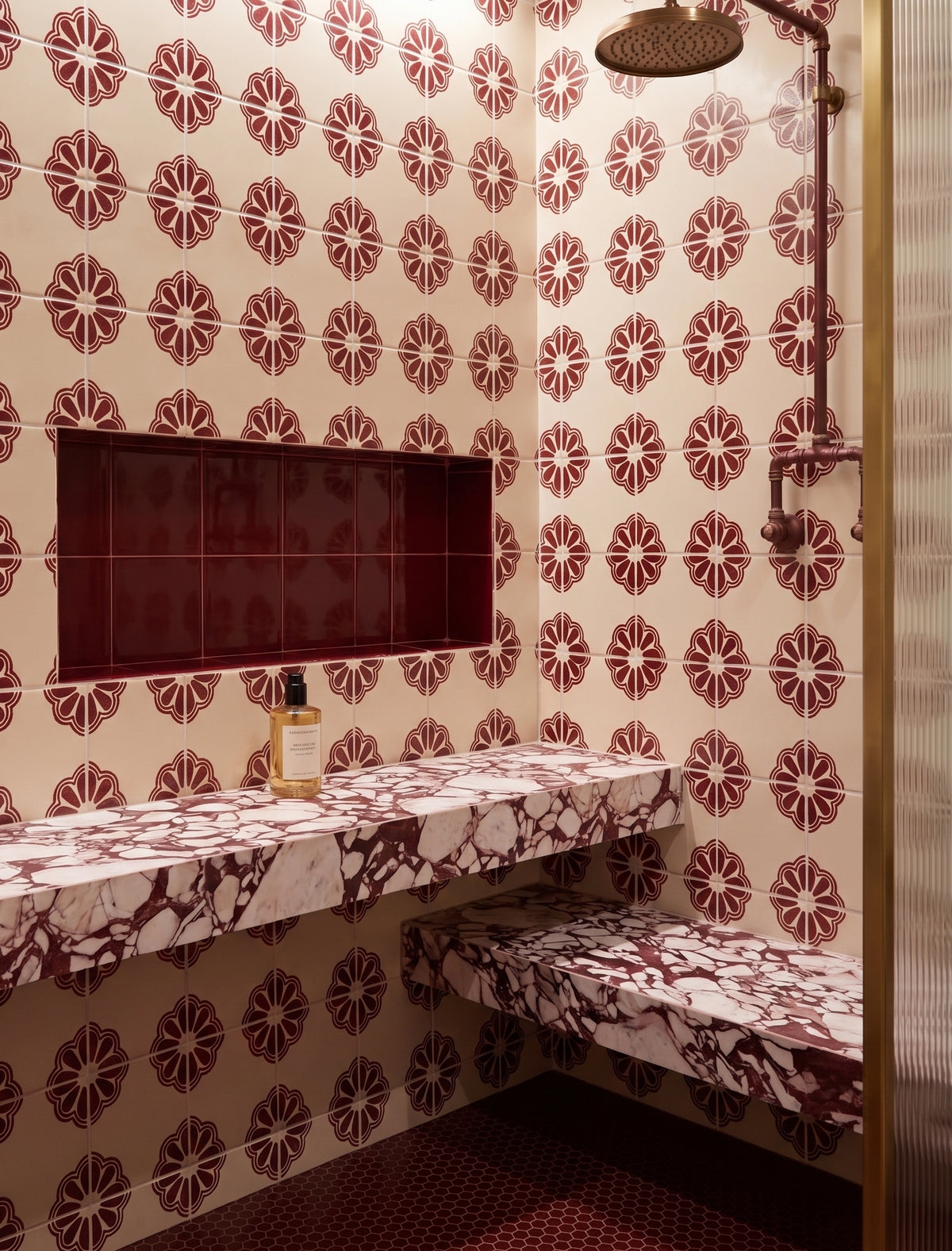 A bathroom interior with repeating circular floral tile pattern in dark red and cream, a built-in stone vanity shelf, matching bench, wall niche, shower fixture, and dark red floor tiles.