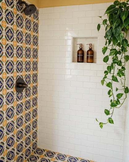 A modern shower features white subway tile walls, a built-in shampoo niche holding amber soap dispensers, vibrant blue and yellow patterned floor and accent tiles, and a cascading trailing pothos plant.