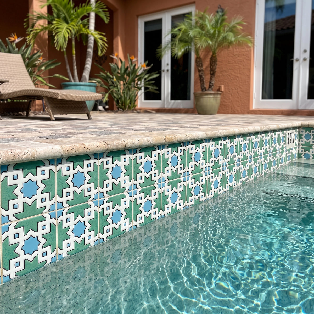 A close-up view of a swimming pool edge decorated with teal, light blue, and white geometric mosaic tiles, set in a backyard with a stone deck, lounge chair, and potted palm trees.