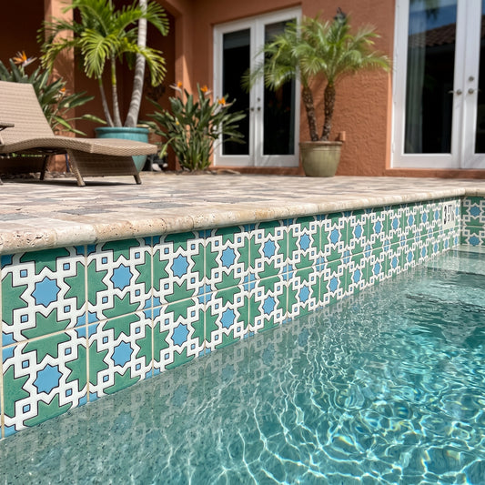 A close-up view of a swimming pool edge decorated with teal, light blue, and white geometric mosaic tiles, set in a backyard with a stone deck, lounge chair, and potted palm trees.