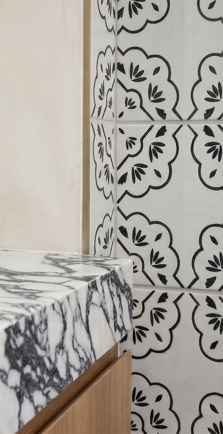 White and black hand-painted tile by Clay Imports, featuring a bold scalloped and floral design. A veined marble countertop and wood cabinet are also shown.