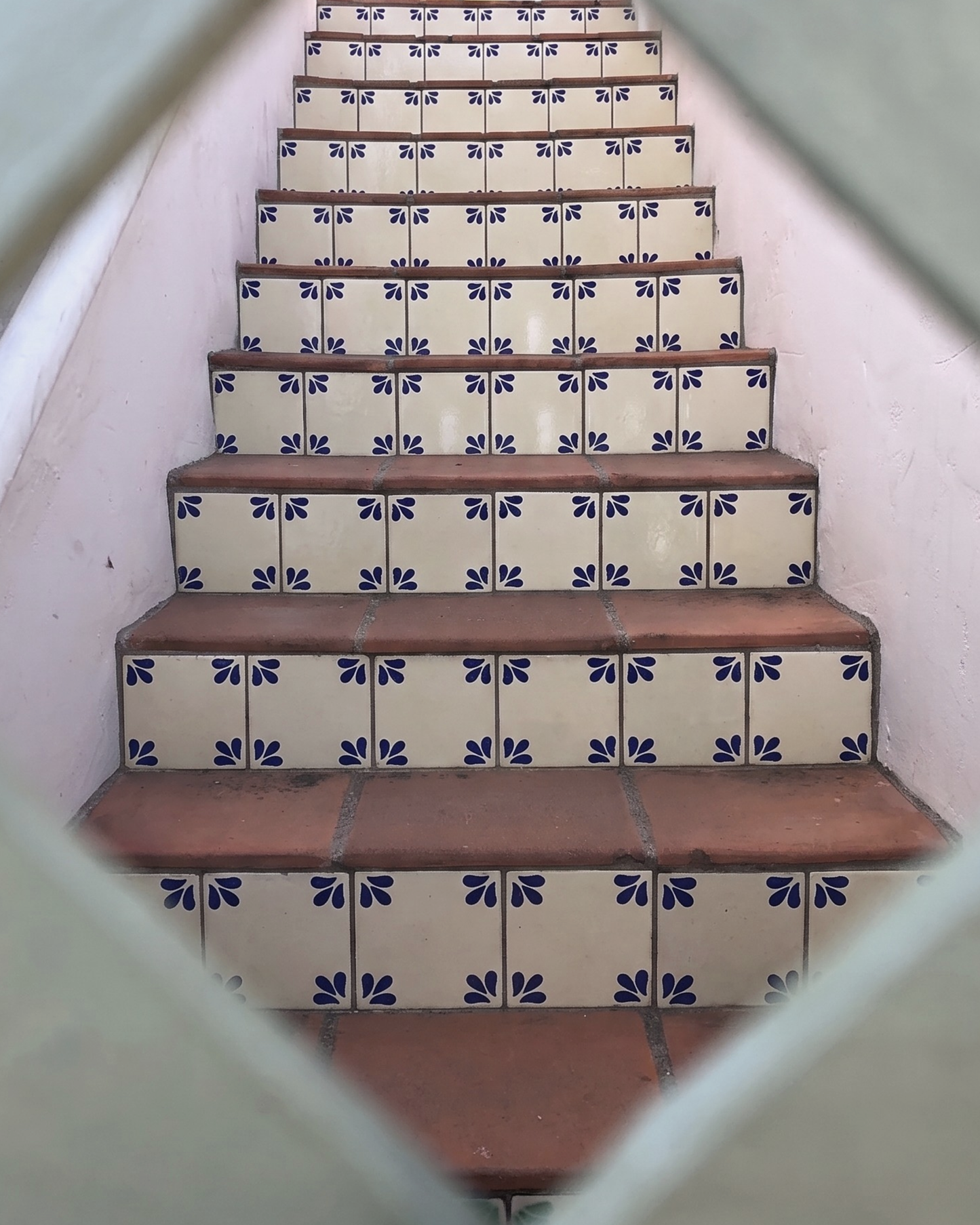A staircase with cream-colored tiles decorated with a blue floral pattern, featuring terracotta-colored steps, viewed through a diamond-shaped opening.