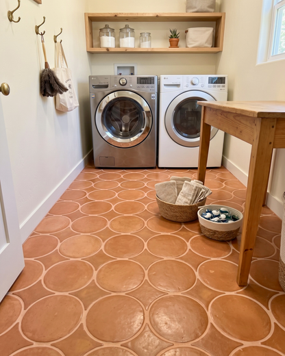 A rustic laundry room featuring terracotta tiled floors with circular patterns, a stainless steel front-load washer, a white front-load dryer, a wooden floating shelf with glass canisters, and a wooden table in the foreground.