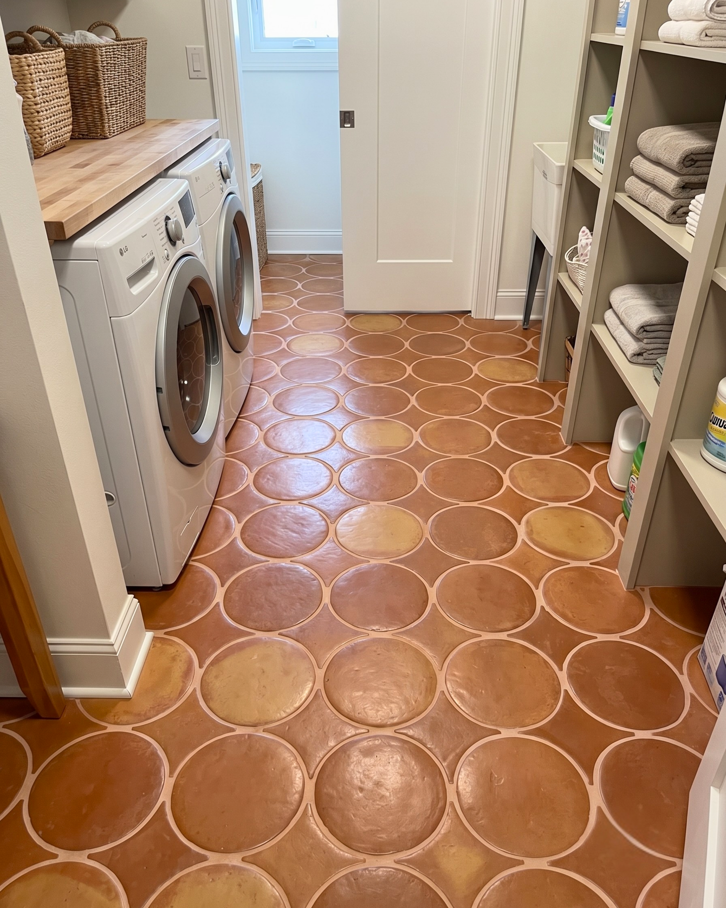 A home laundry room with terracotta-colored circular patterned floor tiles, a white front-loading washing machine, a wooden countertop with woven baskets, and light wood shelving stocked with folded towels and laundry supplies.