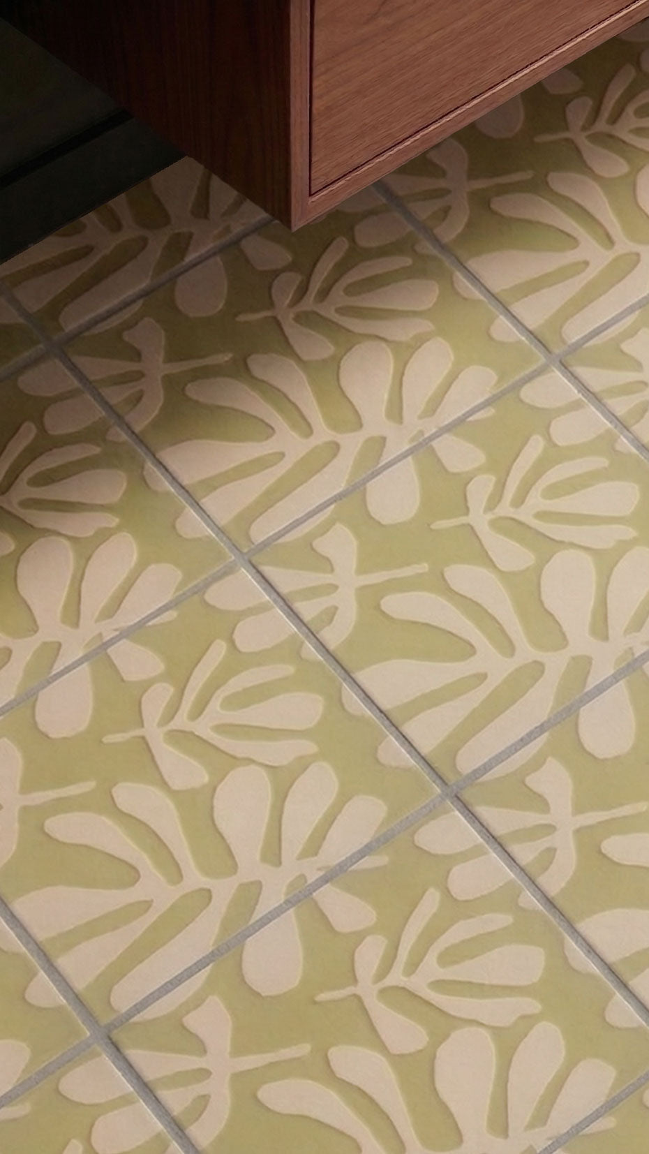 Avocado green pattern tile by Clay Imports featuring off-white floral leaf designs and grey grout lines, below a wooden cabinet.