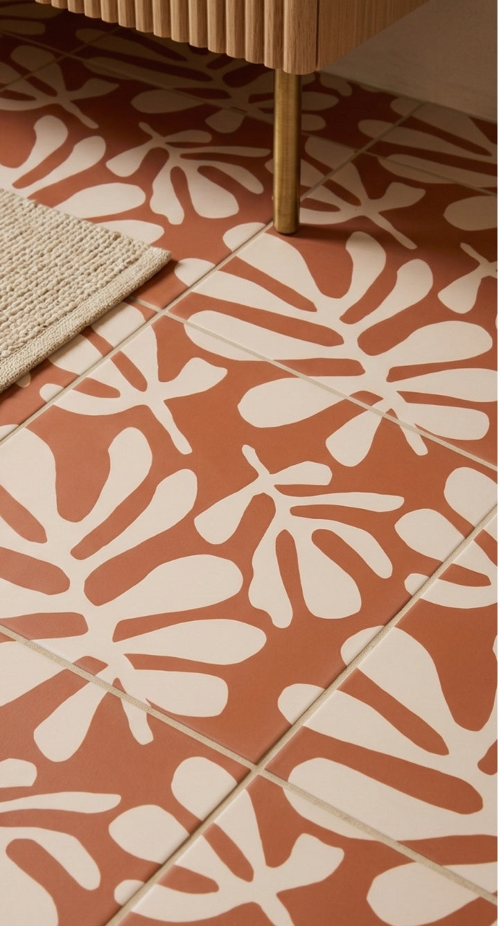 Rust and cream patterned floor tile by Clay Imports, featuring abstract botanical shapes, beside a rug and wooden cabinet leg.