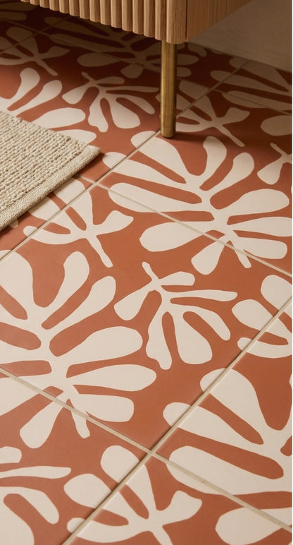 Rust and cream patterned floor tile by Clay Imports, featuring abstract botanical shapes, beside a rug and wooden cabinet leg.