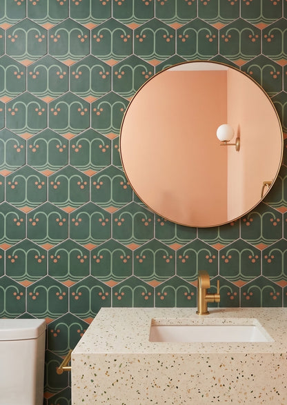 A round gold-rimmed mirror hangs on a patterned green hexagonal tile wall above a speckled terrazzo vanity with a modern brass faucet.