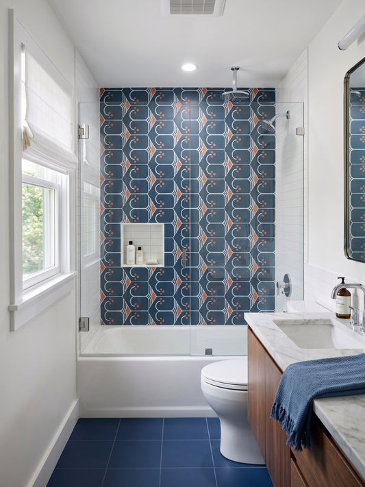 A modern bathroom interior featuring a bold patterned blue and orange tile wall behind a white bathtub, with blue floor tiles, a white toilet, a wood vanity, and a marble countertop.