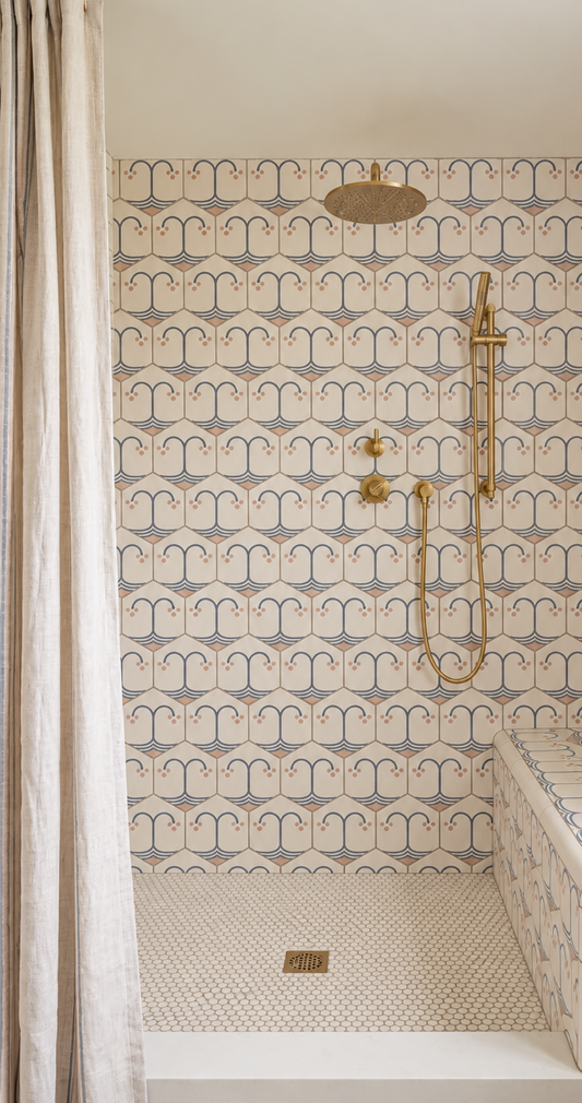A bathroom shower area featuring a wall and floor covered in repeating decorative tiles with a blue and pink motif on a warm cream background. A brass shower fixture and round brass shower head are mounted on the wall, with a light beige curtain visible at the left edge.