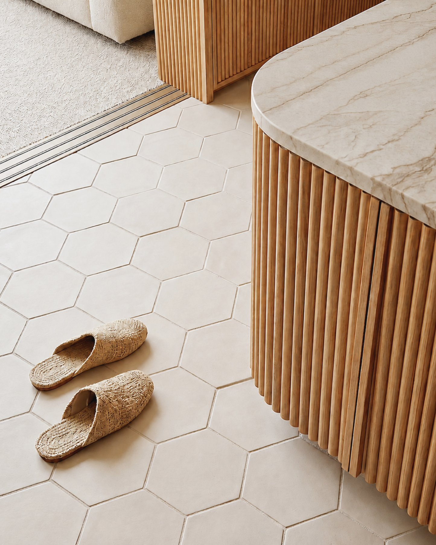 Natural woven jute slip-on slippers placed on a light beige hexagonal tile floor next to a modern wooden slatted cabinet with a marble countertop.