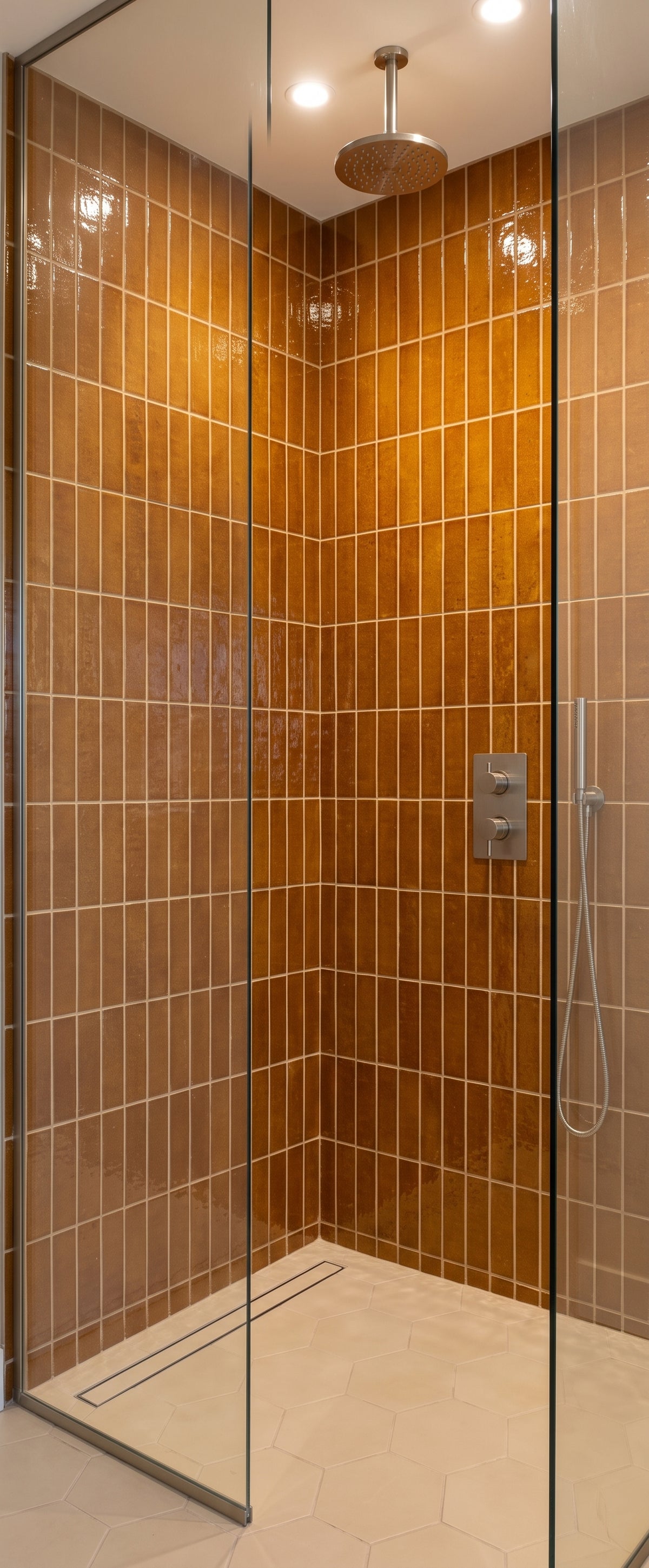 A modern walk-in shower features glossy, amber-colored vertical subway tiles, a round overhead rainfall showerhead, and sleek chrome fixtures behind a clear glass door.