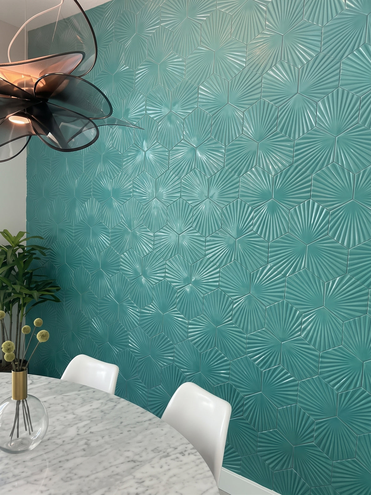 A modern dining room features a striking accent wall covered in textured teal hexagonal tiles beside a marble table and white chairs.
