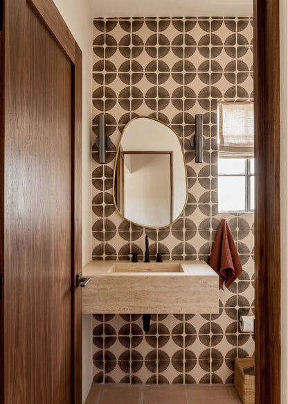 A bathroom interior with a floating stone sink, oval wall mirror, patterned brown and beige tile wall, dark wood door frame, and a rust-colored towel hanging by a window.
