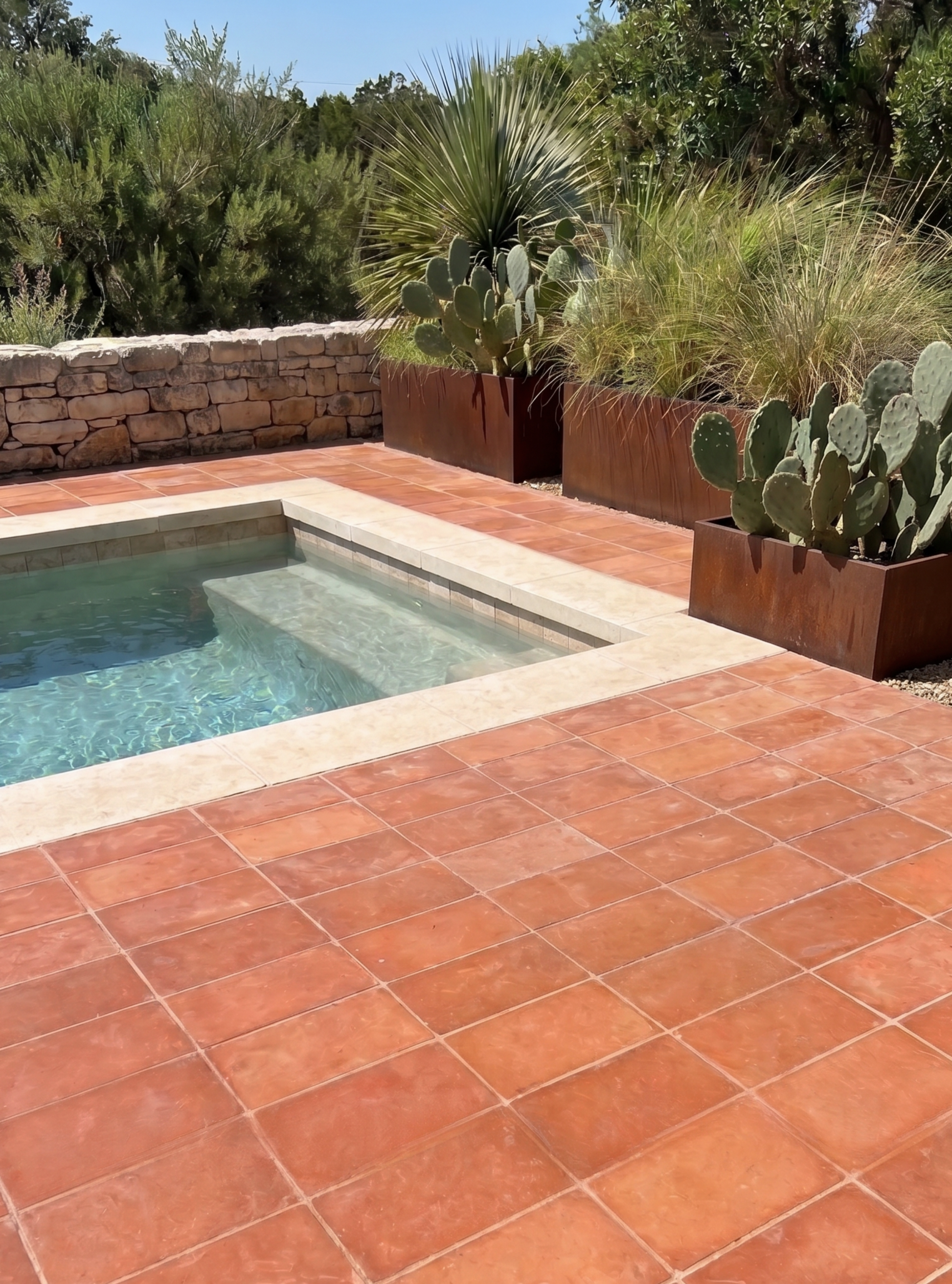 Terracotta-colored rectangular pavers arranged around a small outdoor plunge pool, with a stone wall, cacti, and tall grasses in the background.