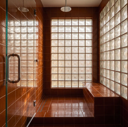 A modern bathroom interior with terracotta brown tiled walls and floor, a built-in bench, a glass shower door with a dark metal handle, and frosted glass block windows. A round ceiling shower head is visible at the top.