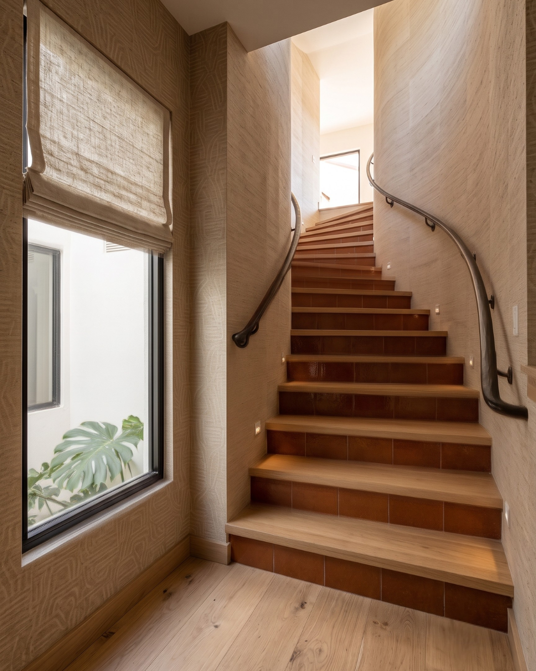 A modern wooden staircase with warm brown steps and matching tiled risers, featuring dark metal railings and beige textured walls. A large window on the left shows a green plant outside, and the staircase leads upward to a bright upper level.