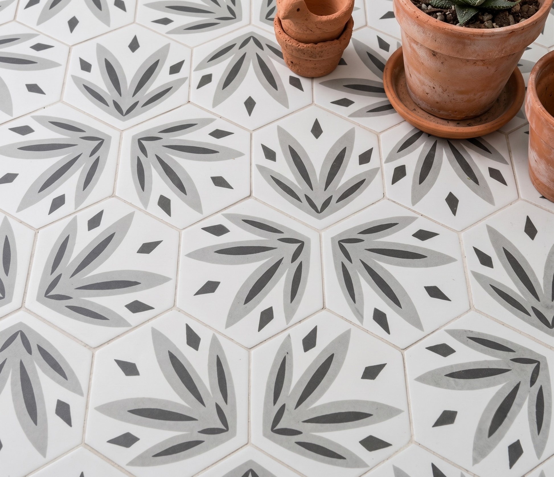 Hexagonal ceramic floor tiles with a repeating gray and charcoal botanical leaf pattern on a white background, styled with terracotta pots in the top right corner.