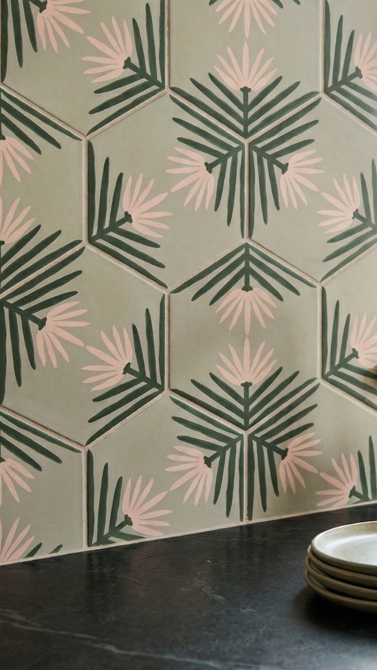 Sage green hexagonal Pattern Tile by Clay Imports, featuring dark green leaves and light pink petals in a repeating botanical pattern, shown above a dark countertop with stacked plates.