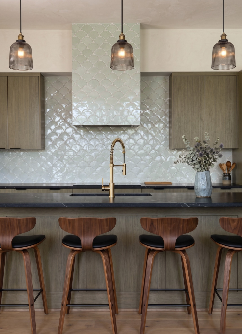 A modern kitchen interior with a scalloped light green tile backsplash, dark stone countertops, three hanging pendant lights, wooden cabinetry, a central island, and four wooden bar stools with black seats.