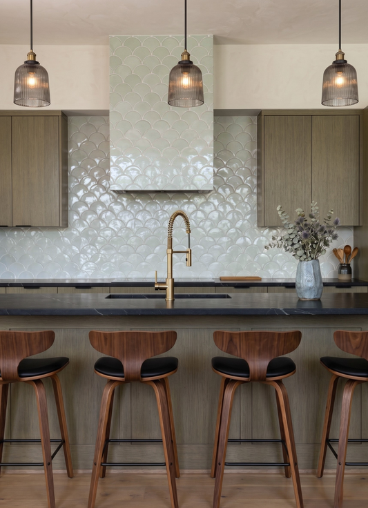 A modern kitchen interior with a scalloped light green tile backsplash, dark stone countertops, three hanging pendant lights, wooden cabinetry, a central island, and four wooden bar stools with black seats.