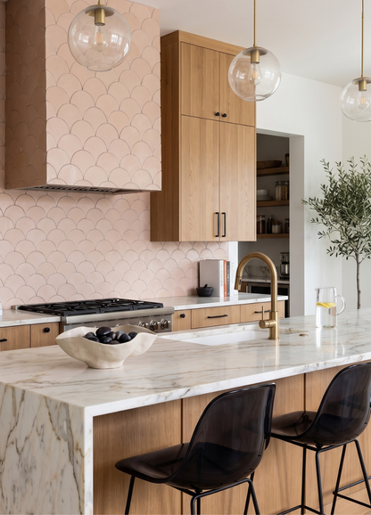 A modern kitchen interior featuring light oak wood cabinets, a pink scallop-patterned tile backsplash, a white marble island countertop, and two black mesh bar stools.