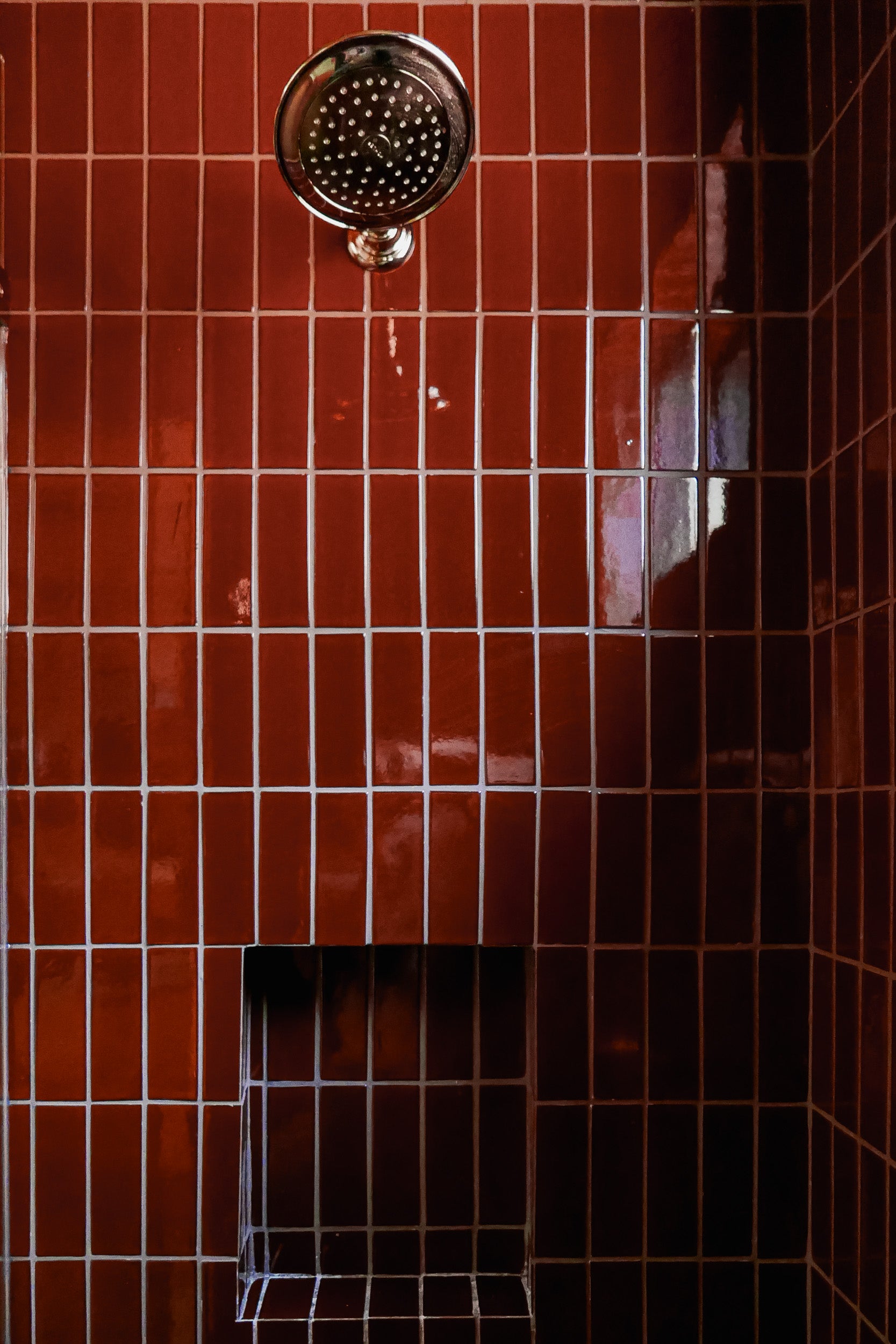 A close-up of a shower wall with glossy reddish-brown square tiles, a round chrome shower head near the top, and a recessed shelf near the bottom.