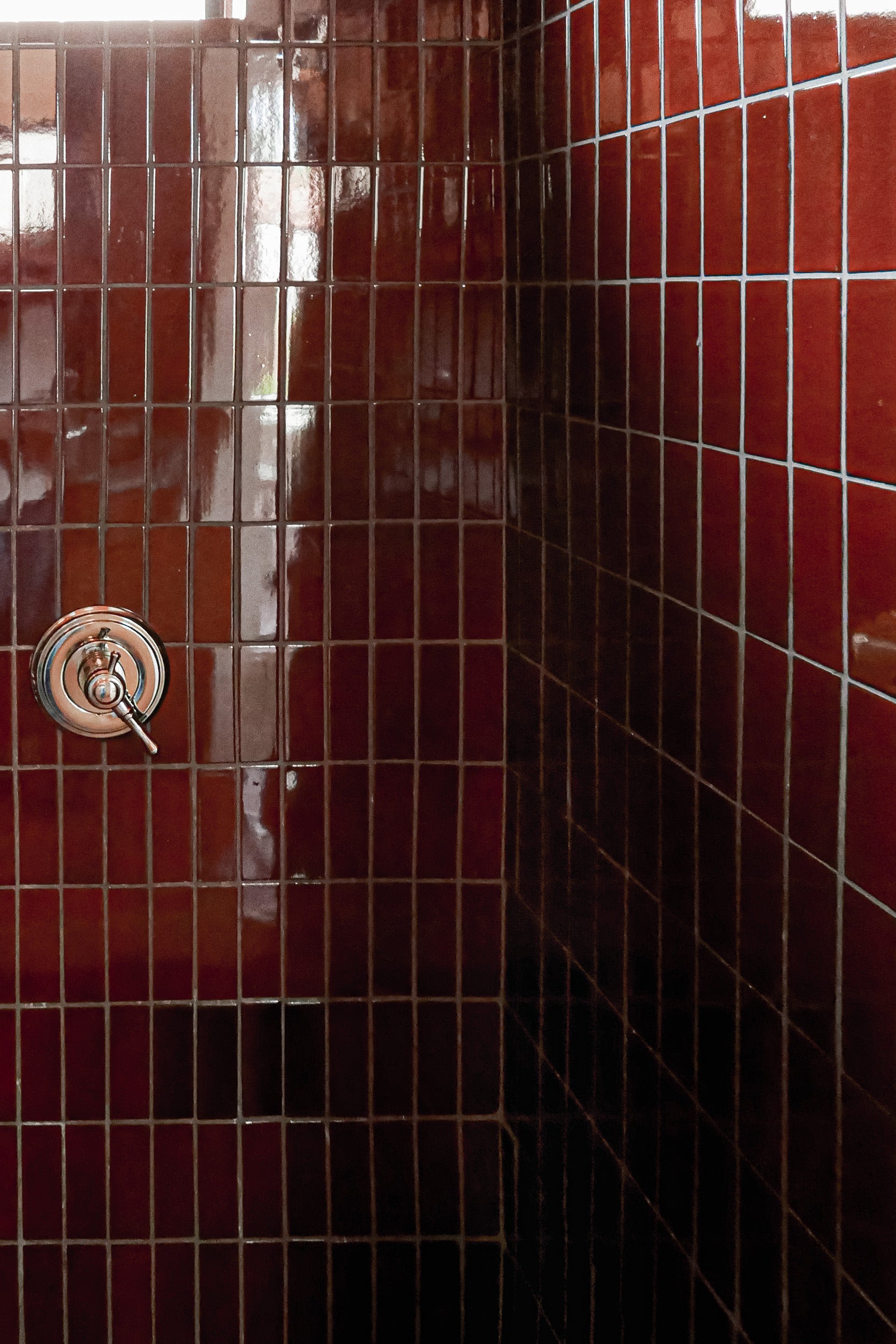 A close-up of a tiled shower corner with glossy dark reddish-brown square tiles and a round metallic shower valve.