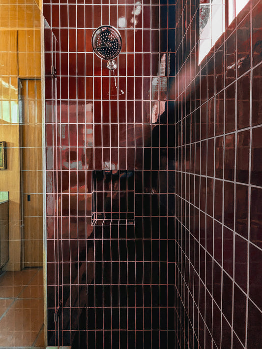 A close-up view of a tiled shower area with glossy dark red-brown wall tiles, a white grout grid pattern, and a round chrome shower head.