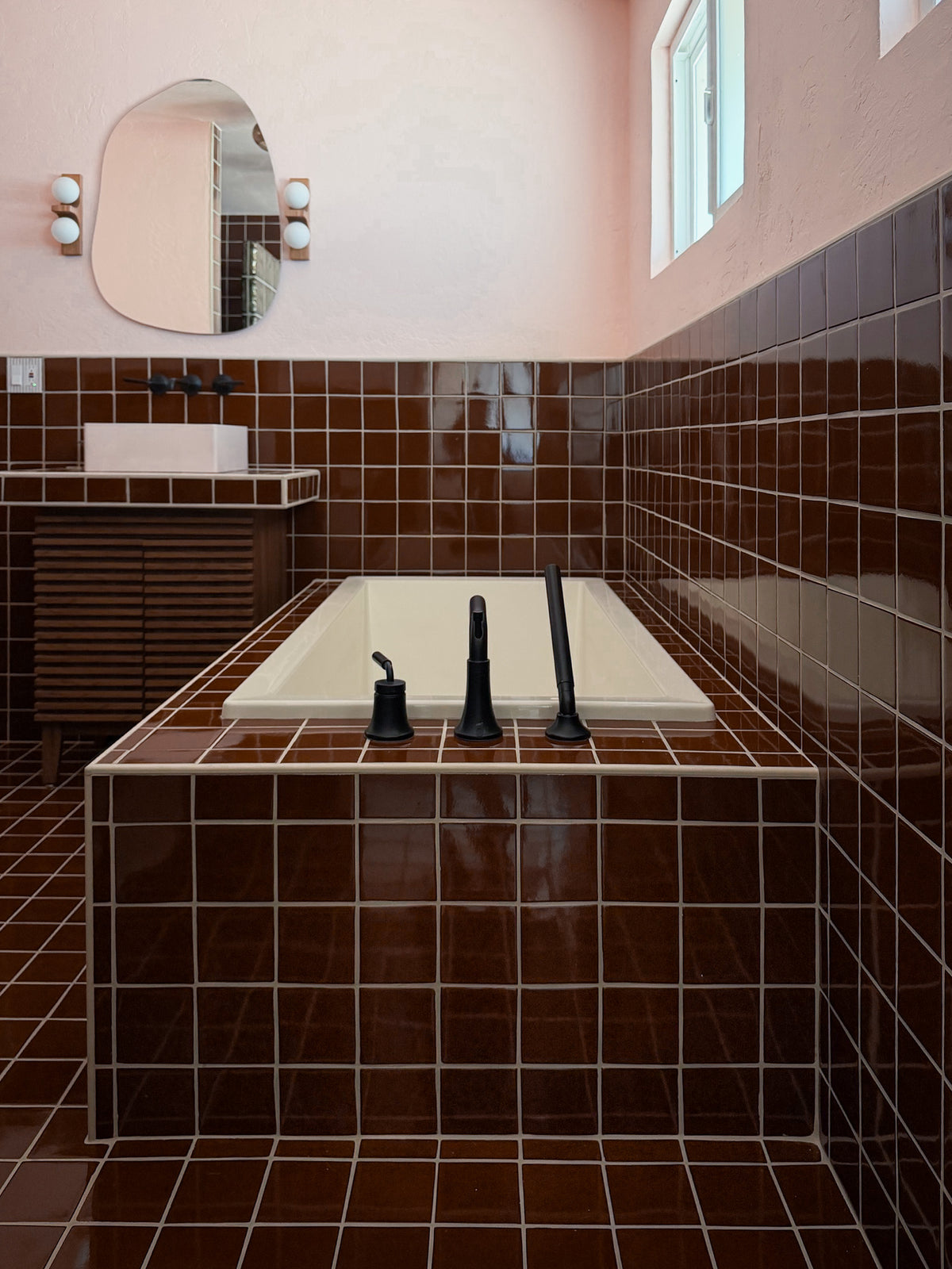 A modern bathroom interior featuring a built-in bathtub with a cream-colored basin and matte black faucet fixtures, surrounded by glossy dark brown square tiles. A matching brown tiled wall and floor surround a vanity with a white rectangular sink and an oval mirror.