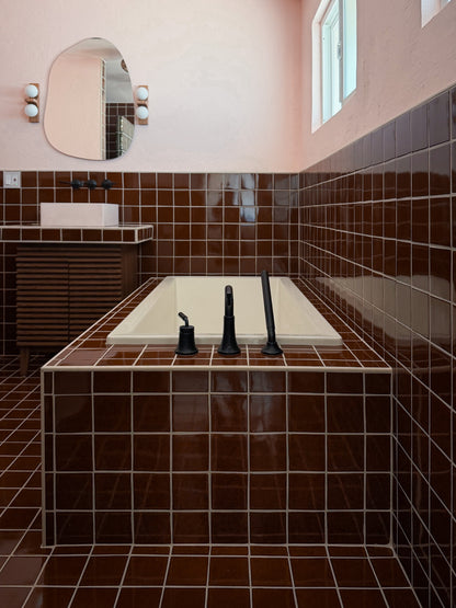 A modern bathroom interior featuring a built-in bathtub with a cream-colored basin and matte black faucet fixtures, surrounded by glossy dark brown square tiles. A matching brown tiled wall and floor surround a vanity with a white rectangular sink and an oval mirror.
