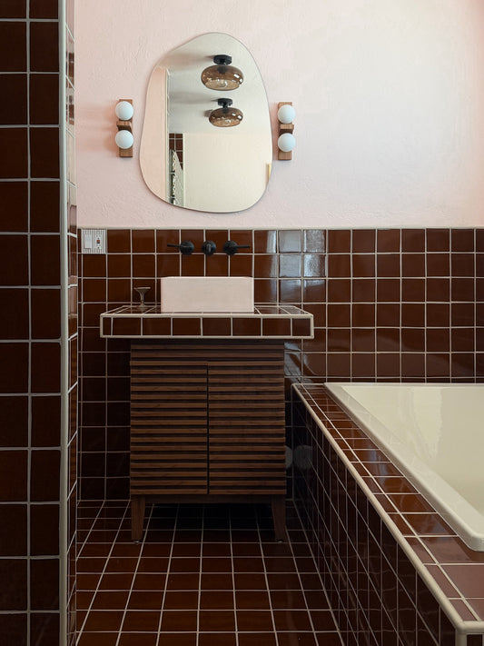 A retro-style bathroom vanity with a dark wood slatted cabinet, a white rectangular sink basin, and a large organic-shaped mirror mounted on a light pink wall above. The bathroom features dark brown square tiles on the floor and wall, a white bathtub on the right, and wall-mounted light fixtures on either side of the mirror.
